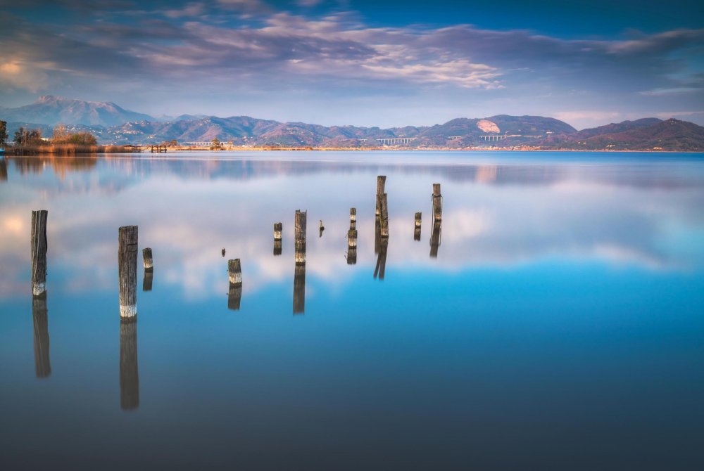 Zonsondergang uitzicht op Massaciuccoli Lake in Torre del Lago. Oude houten palen weerspiegeld in kalm water met Alpi Apuane op de achtergrond, Versilia, Toscane, Italië